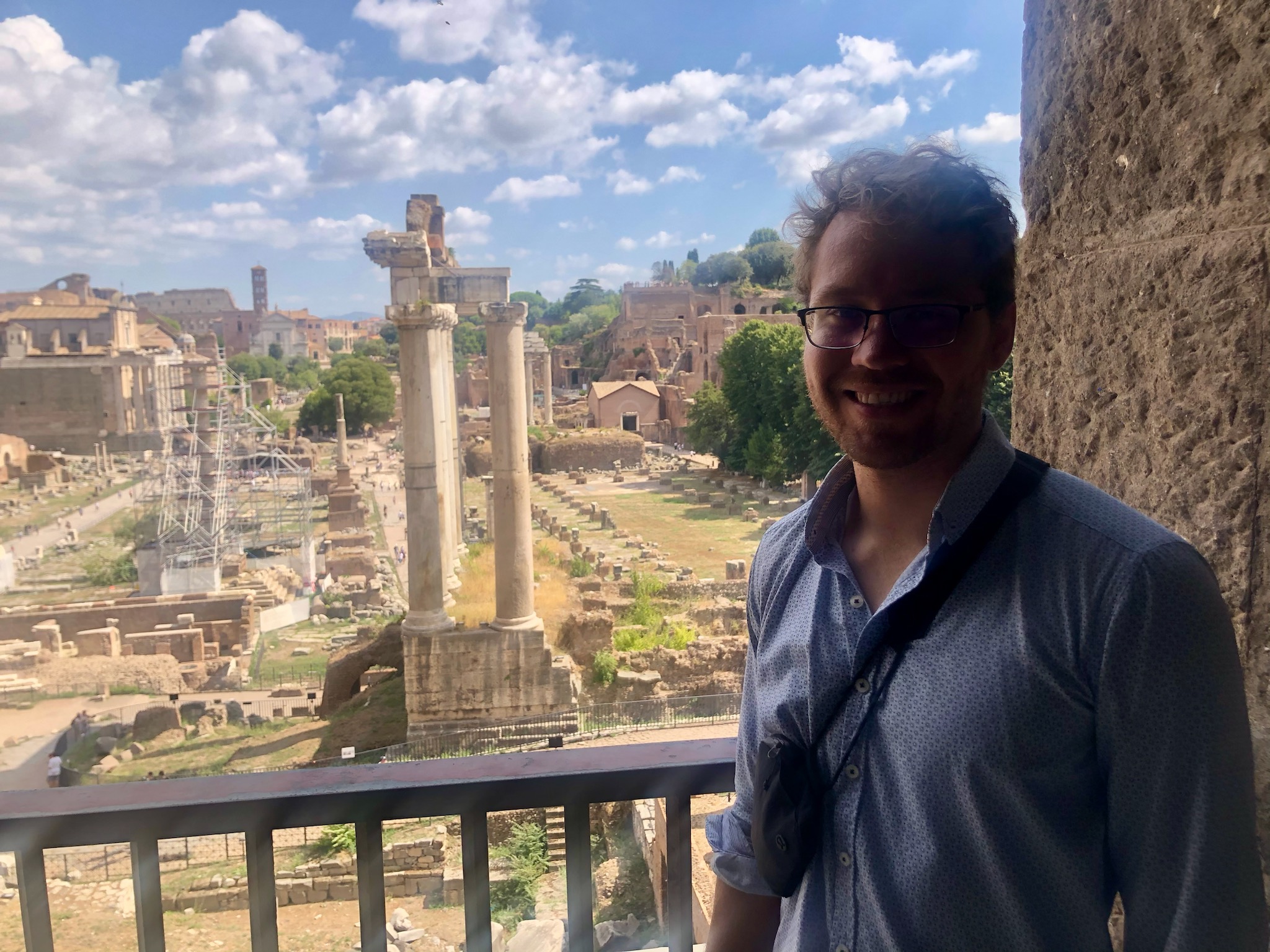 man with glasses standing in front of ancient Greek or Roman ruins