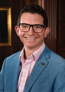 man in blue suit jacket, pink shirt, and glasses in front of a wood wall