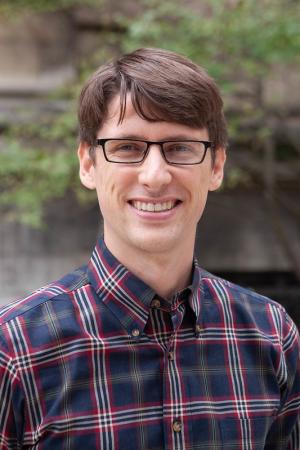 man in a plaid shirt with glasses standing in front of a building with vines