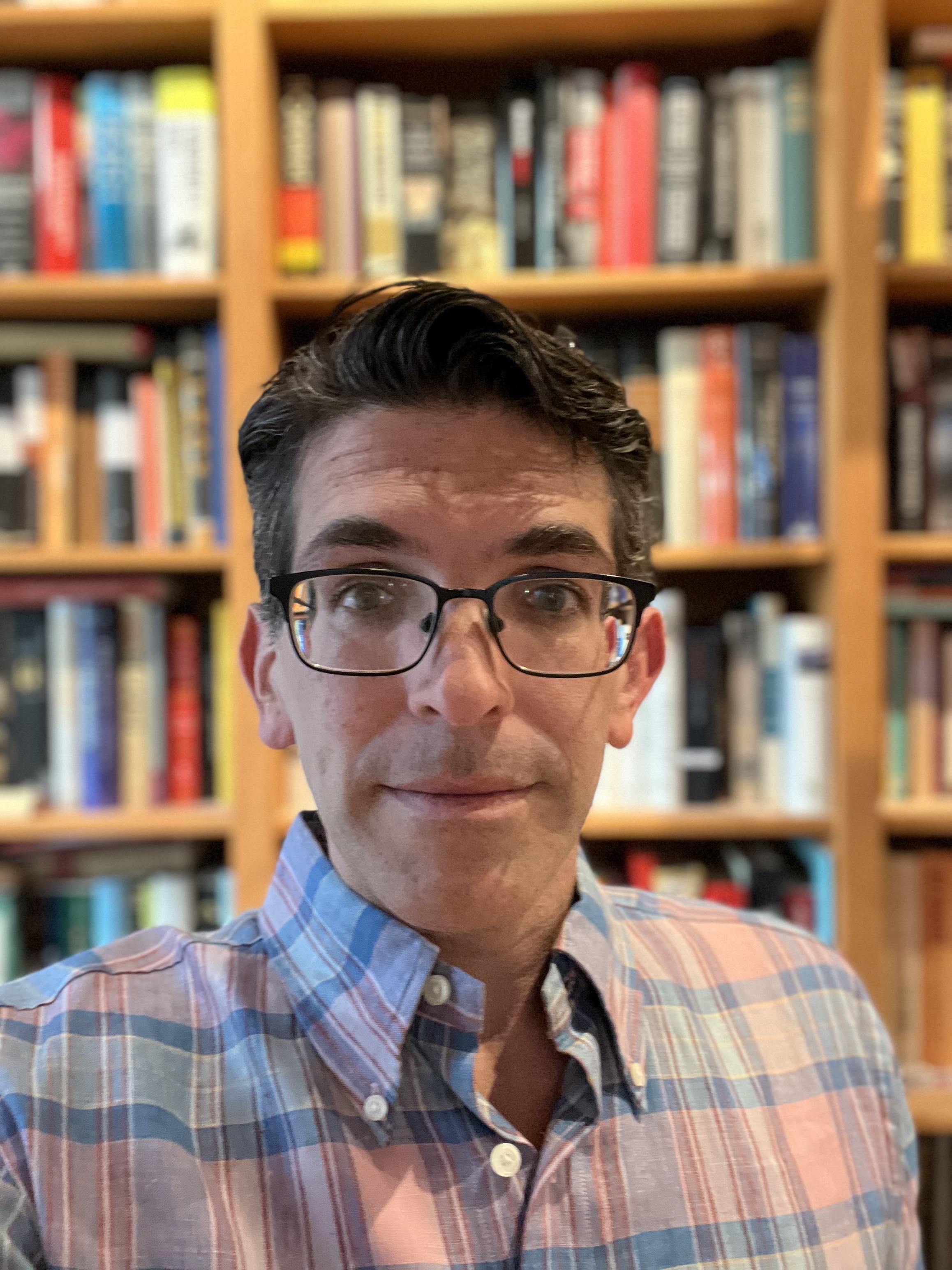 man in plaid shirt, wearing glasses and sitting in front of a bookshelf