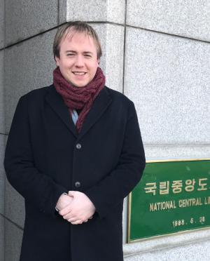 man in a dark coat and maroon scarf standing in front of a building with a green placard