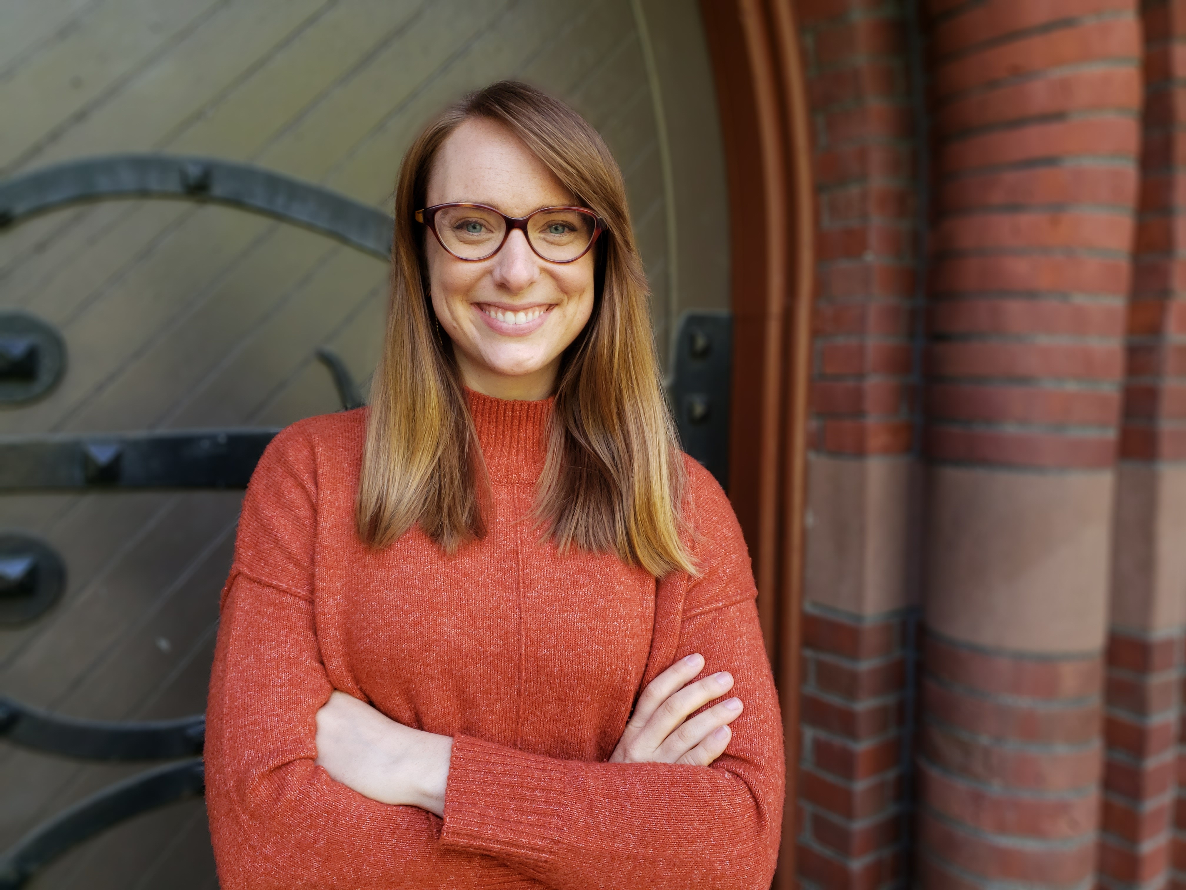 Woman in orange sweater with long hair and wearing glasses. She is standing in front of an arched doorway