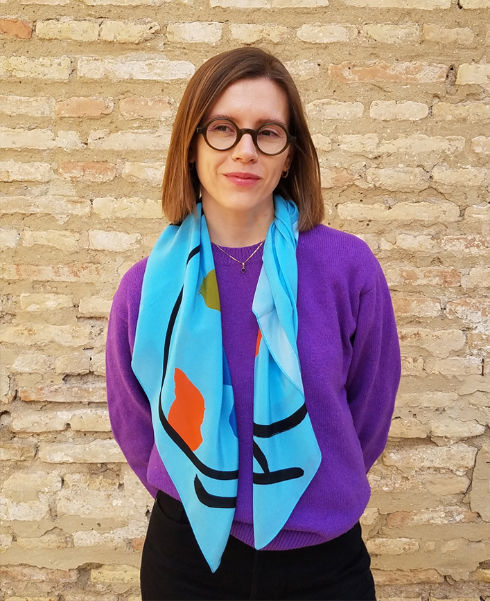 Woman with shoulder-length hair and glasses, wearing a purple sweater and colorful scarf is standing in front of a brick wall