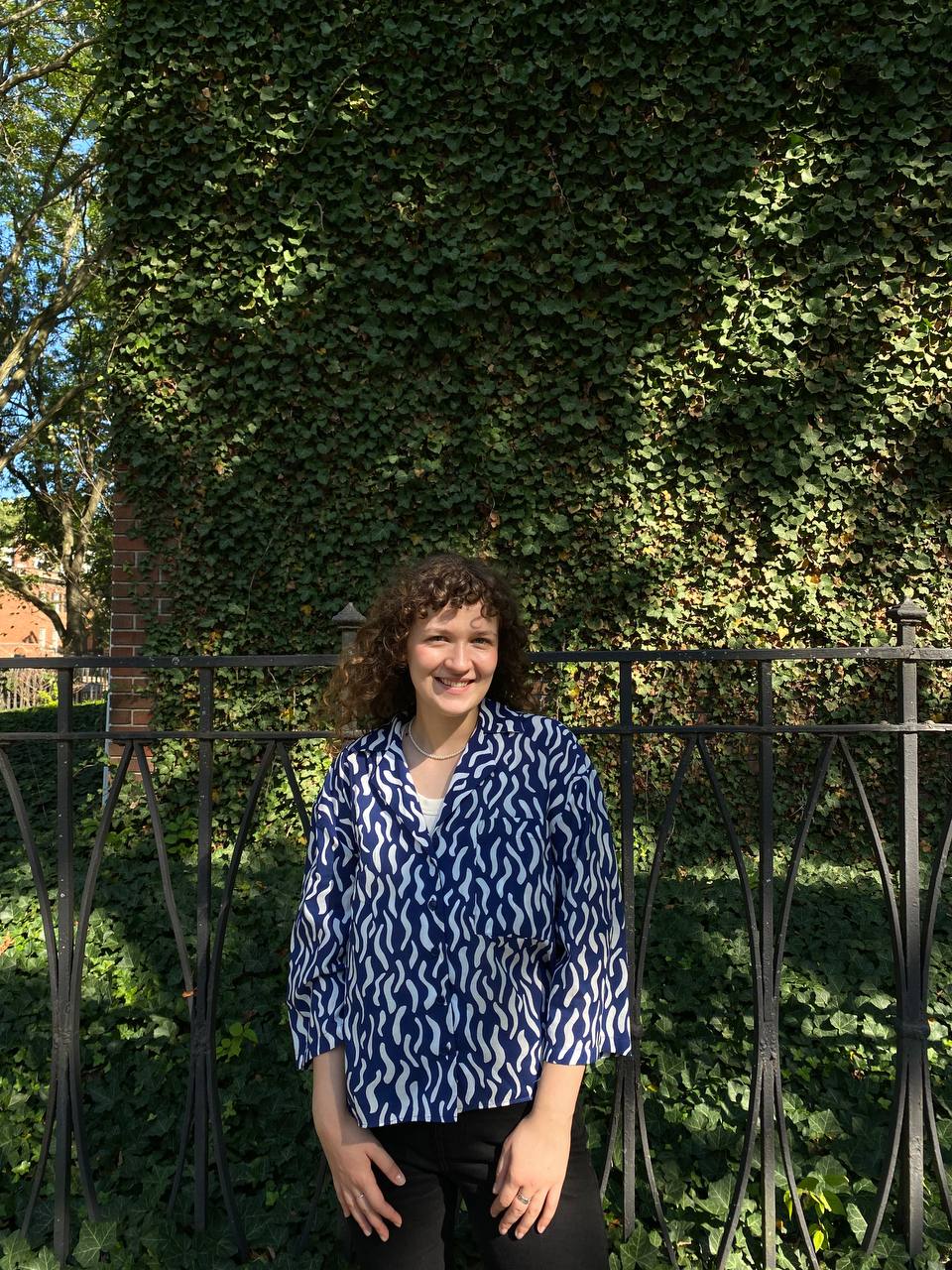 Woman with curly hair wearing a blue and white shirt in front of trees and a fence