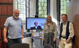 three people and another on zoom standing in a room with windows and books in the background