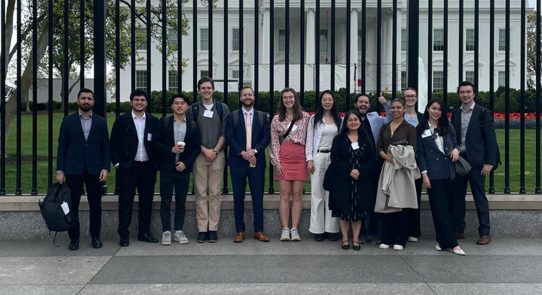 A group of students stand in front of a large black fence with the north face of the US White House in the background.