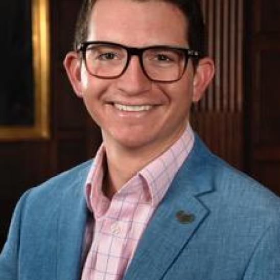 man in blue suit jacket, pink shirt, and glasses in front of a wood wall