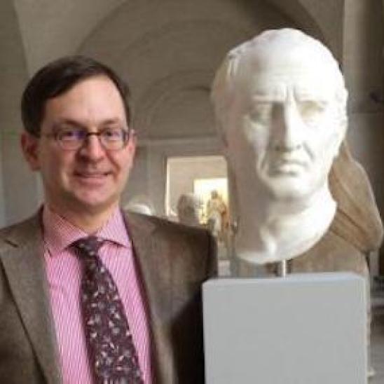 Man wearing round glasses and a suit and tie standing next to a Roman bust
