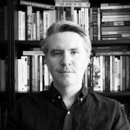 black and white photo of a man in a dark shirt sitting in front of bookshelves