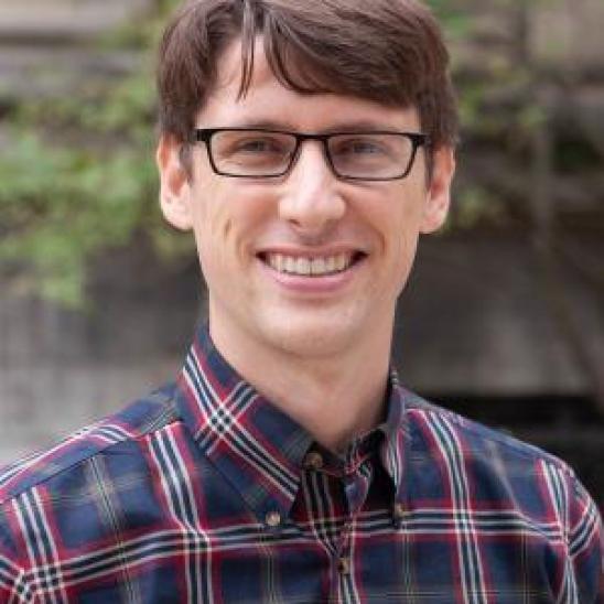 man in a plaid shirt with glasses standing in front of a building with vines