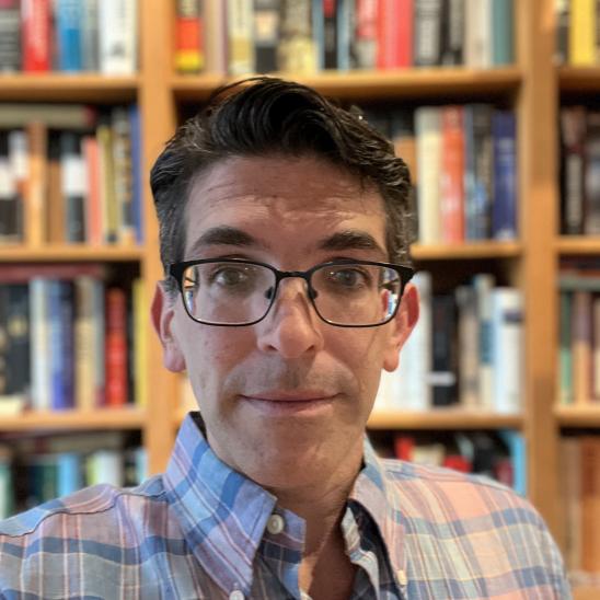 man in plaid shirt, wearing glasses and sitting in front of a bookshelf