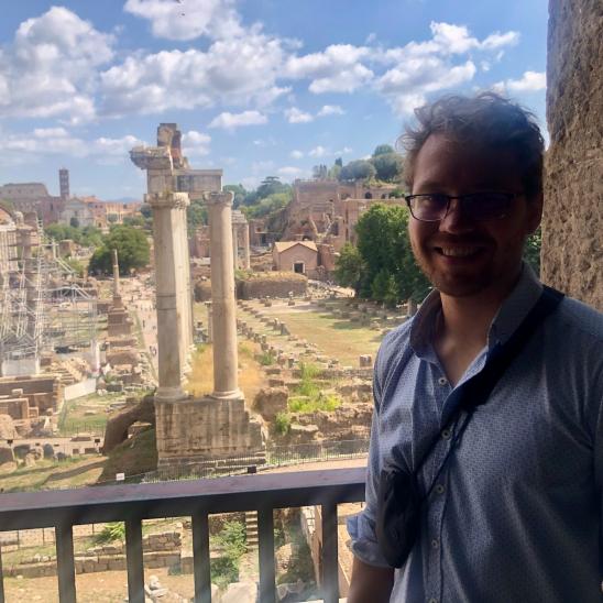man with glasses standing in front of ancient Greek or Roman ruins
