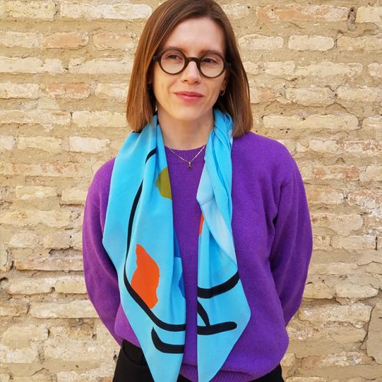 Woman with shoulder-length hair and glasses, wearing a purple sweater and colorful scarf is standing in front of a brick wall