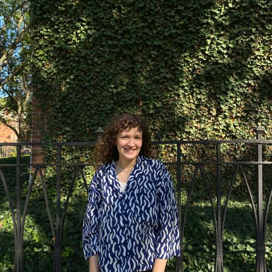 Woman with curly hair wearing a blue and white shirt in front of trees and a fence