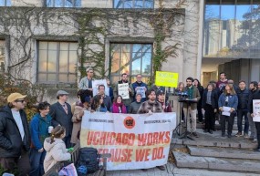 Speakers at a union rally displaying a sign that reads "UChicago Works Because We Do."
